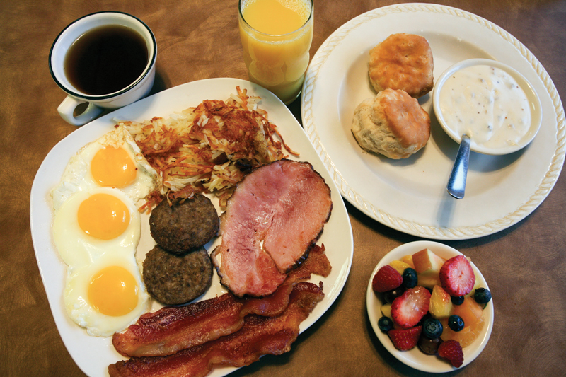 a breakfast of ham, sausage patties, hash browns, sunny-side up eggs, fruit, and biscuits and gravy