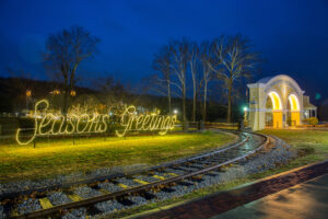 Railroads and a train stop with a decoration sitting in the middle of a well-kept lawn next to the rail roads which reads "Seasons Greetings" in yellow-hued lights.