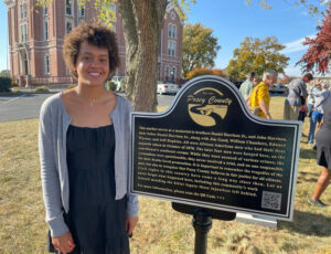 Sophie Kloppenburg stands next to memorial marker, which reads, "This marker serves as a memorial to brothers Daniel Harrison Jr. and John Harrison, their father Daniel Harrison Sr., along with Jim Good, William Chambers, Edward Warner, and Jeff Hopkins. All were African American men who had their lives unjustly taken in October of 1878. The later four men were hung here, on the courthouse's southeast corner. While theyw ere accused of various crimes, the acusations were questionable, they never received a trial, and no one responsible for their deaths faced proescution. It is importat to remember the trageides of the past, but also to recognize that posey county believes in fiar justice for all citizens. Civil rights in this country have come a long way since then. Let us never forget what happened here, including this community's work toward mending the bitter legacy those injustices left behind."